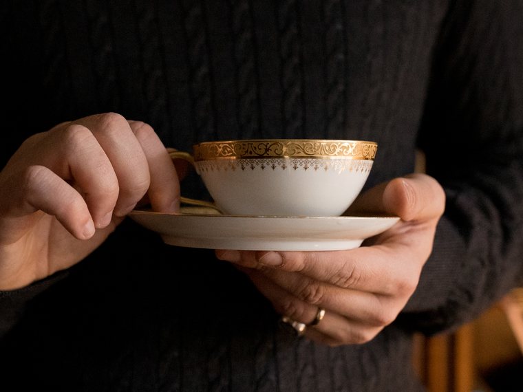 Mãos seguram uma xícara de chá de porcelana branca com borda dourada, sobre o pires, diante de um suéter escuro.